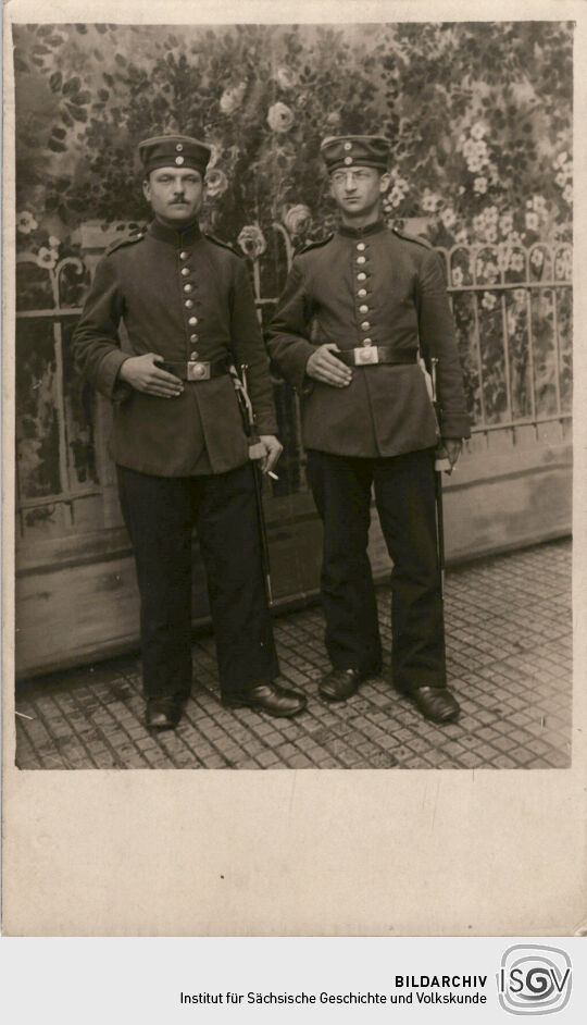 Postkarte: Zwei Soldaten aufgenommen vor einem Hintergrund mit Gartenmotiv