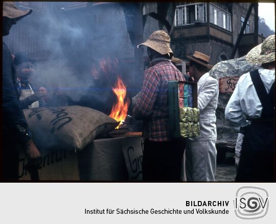 Umzug der Schifferfastnacht in Postelwitz 1959: Teilnehmer an einem Feuer, ein Teilnehmer hat verschiedene Verpackungen (Kaffe) an seinem Rücken befestigt