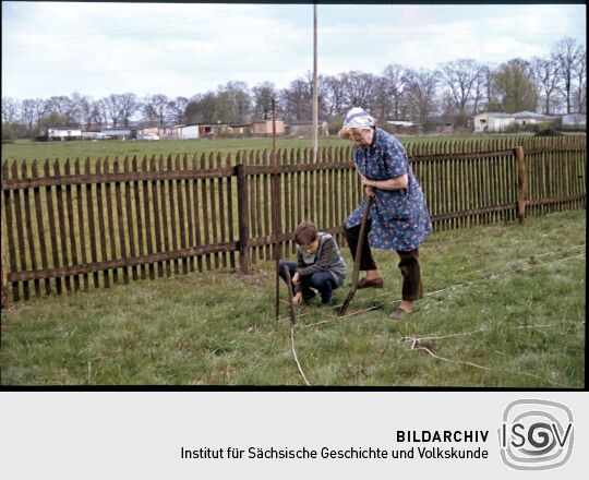 Eine Frau und ein Kind in Kittelschürzen im Kleingarten.