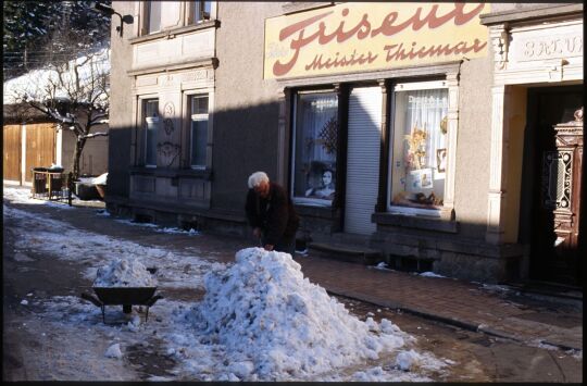 Mann beim Schneeschippen mit einer Schubkarre vor einem Friseursalon in Schmiedeberg