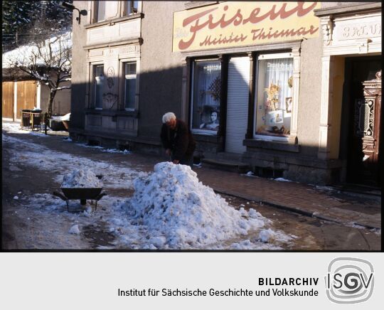 Mann beim Schneeschippen mit einer Schubkarre vor einem Friseursalon in Schmiedeberg