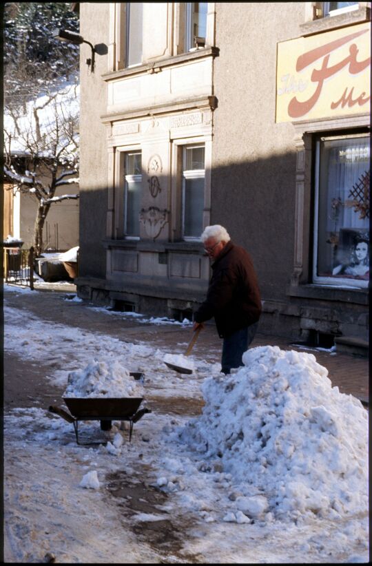 Mann beim Schneeschippen mit einer Schubkarre vor einem Friseursalon in Schmiedeberg