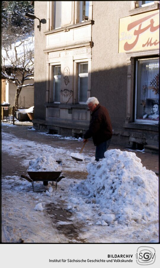 Mann beim Schneeschippen mit einer Schubkarre vor einem Friseursalon in Schmiedeberg